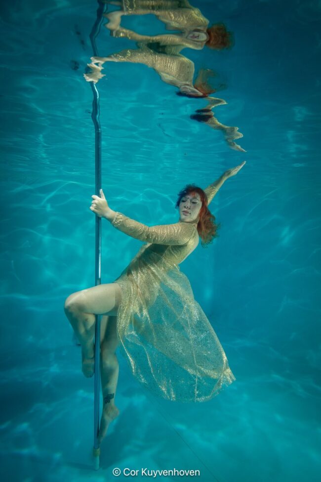 Model met een paaldans paal onder water. Alternatieve onderwater foto van een vrouw in een gouden jurk die balanceert met een paaldans paal in een zwembad.