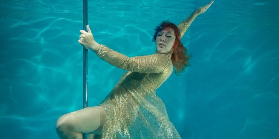 Alternatieve onderwater foto van een vrouw in een gouden jurk die balanceert met een paaldans paal in een zwembad.