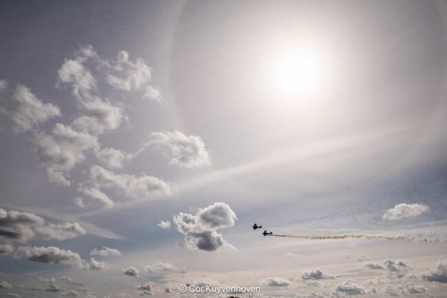 Luchtvaartshow met een vliegtuig dat door de lucht vliegt onder een bewolkte hemel met de zon op de achtergrond.