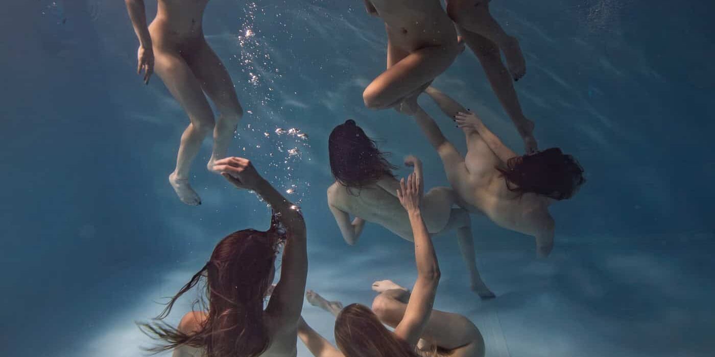 Zeven vrouwen zwemmen onder water, dansend en spelend, in een zwembad met helderblauw water.