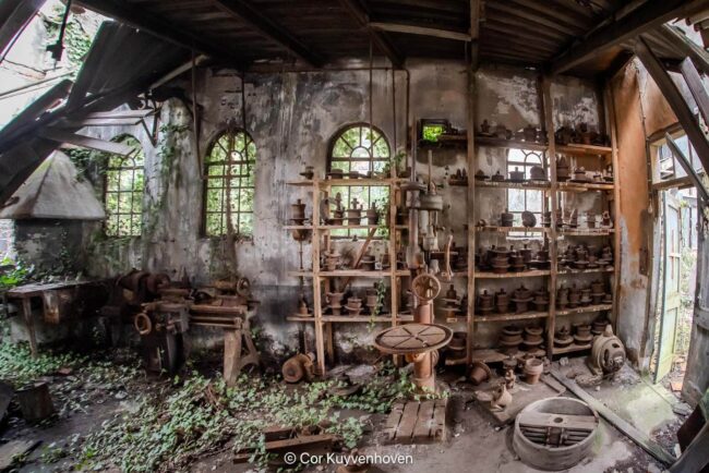 Oude werkplaats met keramische potten, gereedschap en houten rekken in een vervallen, rustieke omgeving.