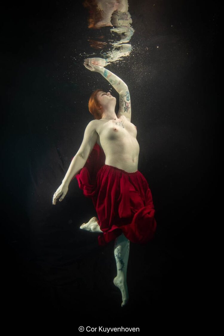 Onderwaterfoto van een vrouw met tattoos die onder water zwemt, gekleed in een rode rok en topless, met haar hoofd omhoog en haar arm gestrekt.