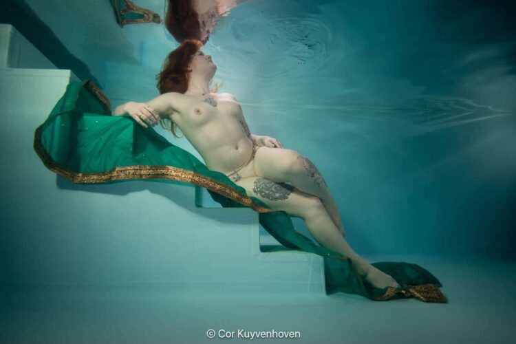 Een vrouw drijft ontspannen in een zwembad, omgeven door turquoise water, met een groene en gouden jurk die haar elegantie benadrukt.