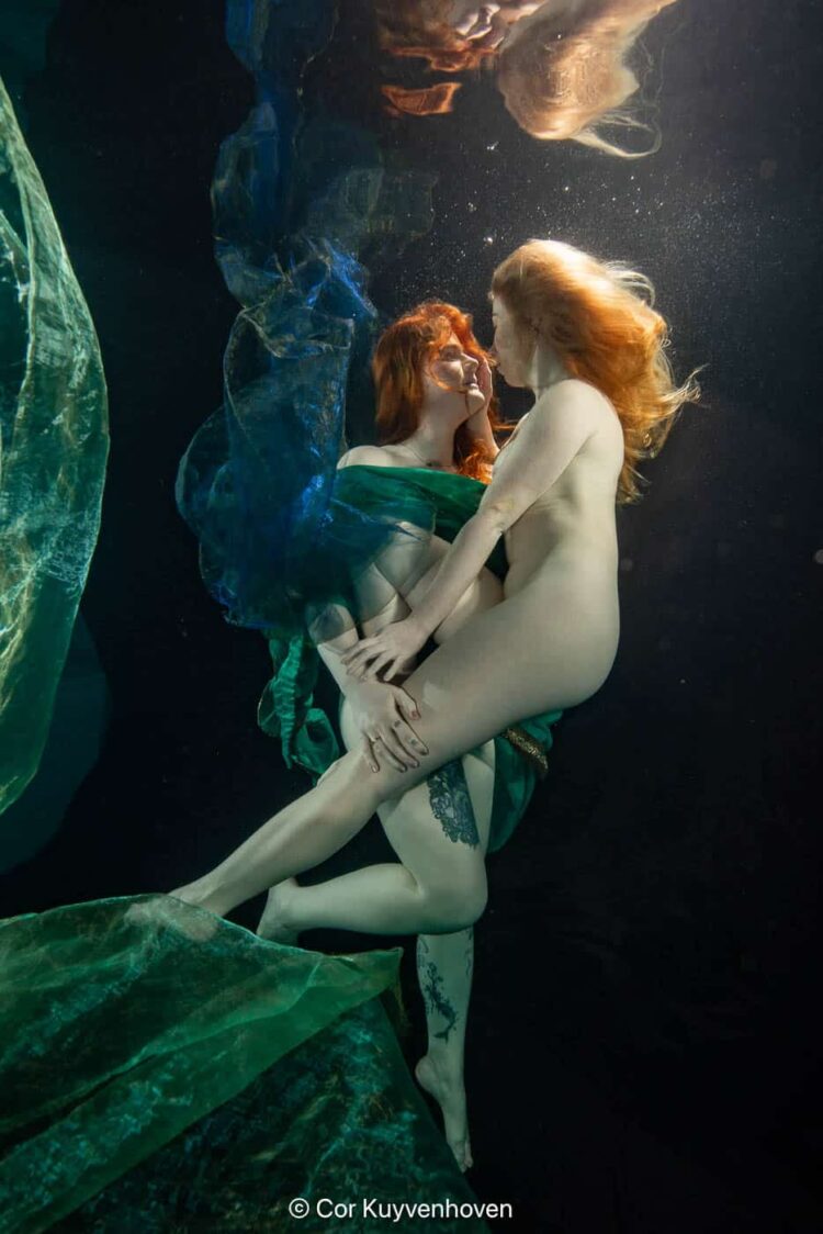 Onderwaterfoto van twee vrouwen die elkaar omarmen, met kleurrijke stoffen en waterbubbels, artistiek en emotioneel vastgelegd.