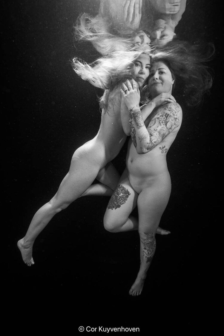 Onderwaterfoto van twee vrouwen met tattoos, die samen drijven in het water, met hun haar en lichamen elegant bewegend.
