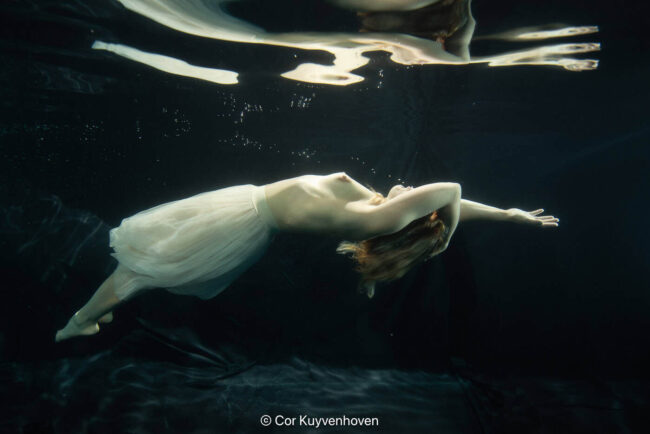 Onderwaterfoto van een vrouw in een witte jurk die horizontaal drijft in het water, met reflecties en golven boven haar.