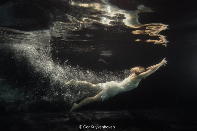 Zwemmer onder water, bewegend door het water met een gestrekte arm, in een donkere omgeving, professionele sportfotografie.