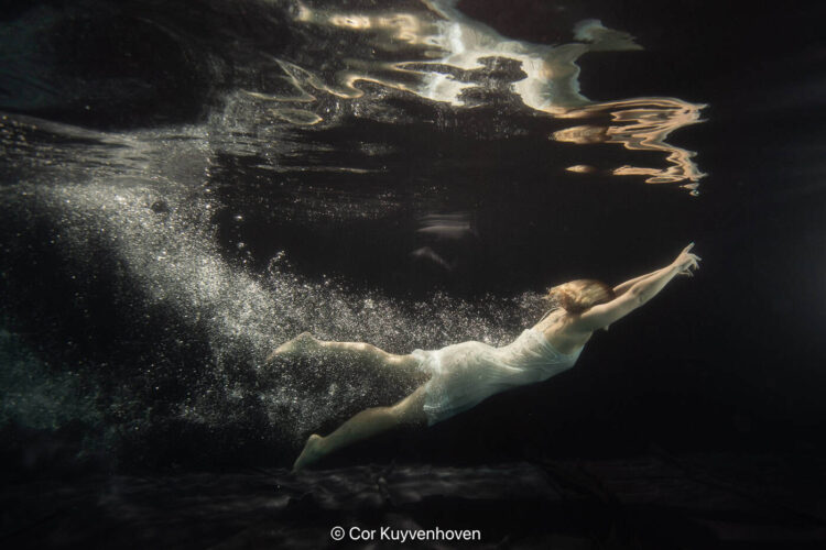 Zwemmer onder water, bewegend door het water met een gestrekte arm, in een donkere omgeving, professionele sportfotografie.