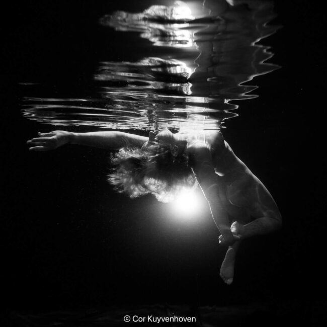 Zwart-wit foto van een naakte vrouw die onder water drijft, met licht dat door het water schijnt, artistiek en rustgevend.