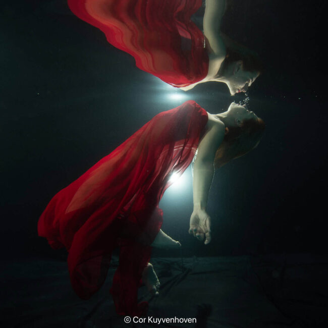 Onderwaterfoto van een vrouw in een rode jurk die onderwater het water oppervlakte aan raakt, met lichtstralen door het water.
