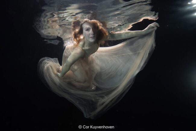 Onderwaterfoto van een vrouw met een lichte jurk die zweeft in het water, met reflecties en lichtstralen.