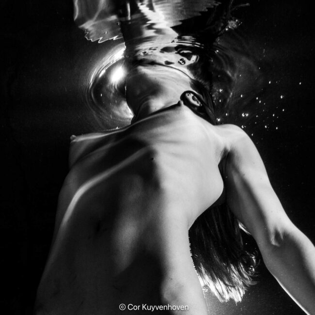 Zwart-wit foto van een vrouw die onder water zweeft, met haar hoofd en haar haar onder water, en haar lichaam zichtbaar vanaf de schouder tot de taille.